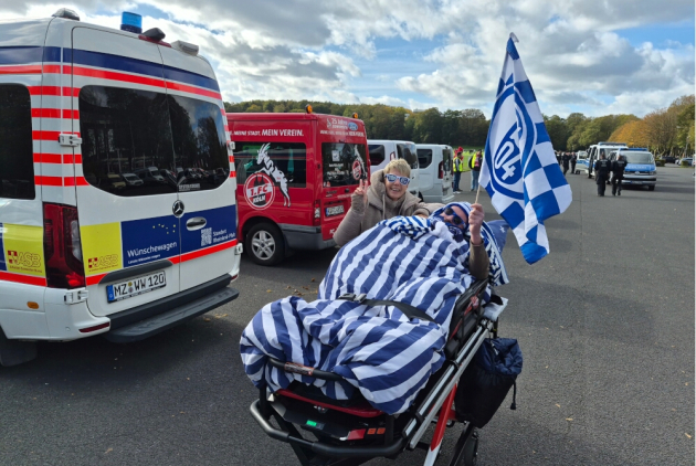 Neuwied: Bewohner mit Schalkeflagge im Bett neben Betreuungsdienstmitarbeiterin auf Stadionparkplatz.